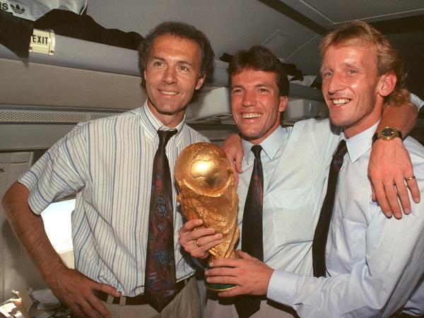 Im Flugzeug beim Rückflug von Rom nach Frankfurt präsentieren DFB-Teamchef Franz Beckenbauer, Lothar Matthäus und Abwehrspieler Andreas Brehme strahlend den zwei Tage zuvor gewonnenen WM-Pokal. 